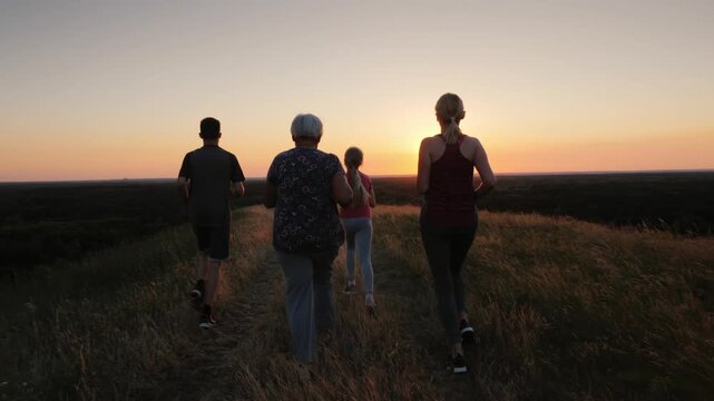 Group of four active family members running together on top of grassy hill during beautiful summer sunset in scenic rural landscape view capturing health and wellness outdoor activity 