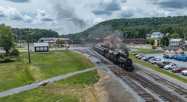 Aerial view of the historic East Broad Top Railroad steam train pulling into the station with smoke, surrounded by rural landscape and historic buildings in Rockhill, Pennsylvania, United States.