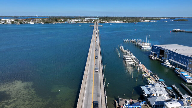 Aerial view of the Anna Maria Island Bridge spanning blue waters with cars driving across, near a marina with boats and a blue building in Bradenton, Florida, United States.