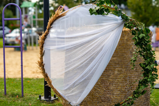 Decorative circular frame featuring draped white tulle, ivy foliage, and dried cereal ears set beside a straw bale outdoors, natural daylight lighting, close-up photograph.   