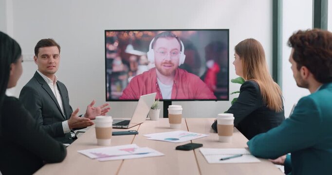 Caucasian executive talks as gestures to online speaker on screen at office call. Diverse business team watches video meeting following discussion attentvely