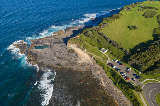 Aerial view of a coastal carpark alongside a rock ledge and green grassy cliffs