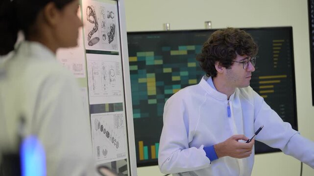 Seated curly-haired engineer with glasses points a pen at a bionic arm held by a colleague in front of a data heat map screen, during a collaborative session, an embodied AI research scene