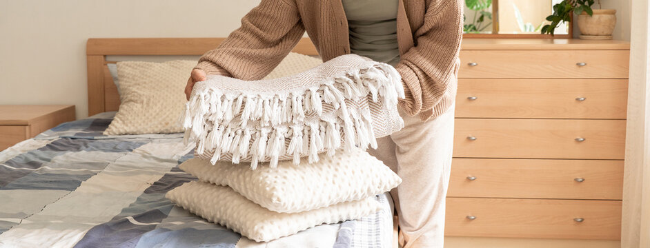 Woman in beige cardigan holds stack of decorative soft pillows and fringed throw blanket over bed with wooden furniture. Slow living, mindfulness, seasonal rituals