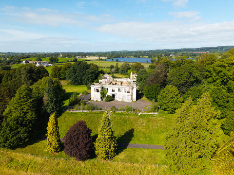 Aerial view of a derelict mansion surrounded by overgrown trees and lush green fields with a lake in the distance in Lisburn, Northern Ireland, United Kingdom.