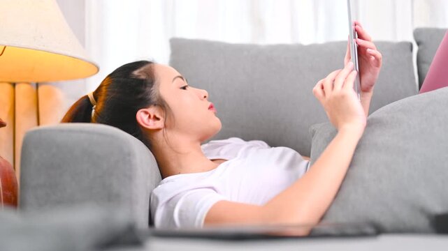 A woman holds a smartphone while wearing black headphones beside a gray sofa. The woman looks at the phone screen in the home interior; the display is too small to identify any app or service.