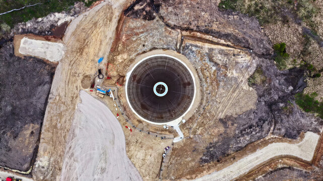 Aerial view of a circular wind turbine foundation under construction with exposed rebar and concrete, surrounded by excavated earth and soil in Latvija.