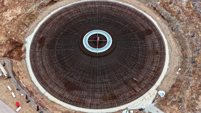 Aerial view of a circular wind turbine foundation under construction, featuring an intricate steel rebar reinforcement mesh and a central metal ring set within an excavated site in Latvia.