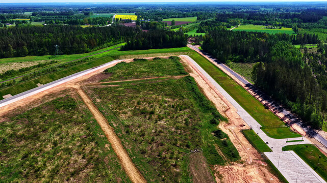 Aerial view of a large cleared plot of land with dirt paths, a paved road, and surrounding forest in the rural landscape of Latvia.