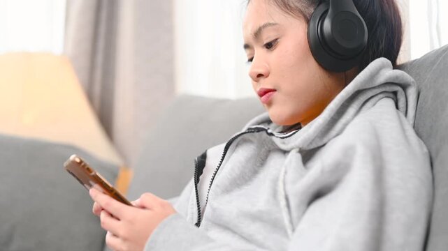 A woman holds a smartphone while wearing black headphones beside a gray sofa. The woman looks at the phone screen in the home interior; the display is too small to identify any app or service.