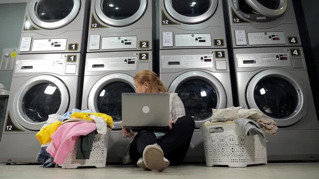 A woman with a laptop is sitting on the laundry room floor against the background of a row of washing machines and dryers, next to baskets of dirty laundry. The woman is waiting for the end of washing
