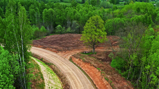Aerial view of a winding dirt road cutting through a lush green forest with a recently cleared patch of land in Latvija.