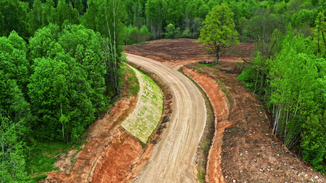 Aerial view of a winding dirt road cutting through a dense green forest with exposed red clay embankments in Latvia.