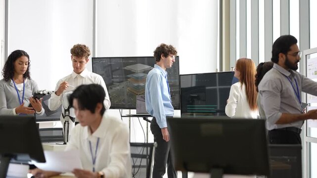 Asian engineer reviews documents at his desk while teammates test a humanoid robot hand and talk at screens in a bright office, during a practical trial, a digital transformation concept
