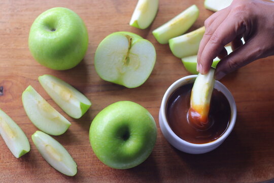 Green apple wedge dipped into caramel sauce with fresh apple slices on a wooden board
