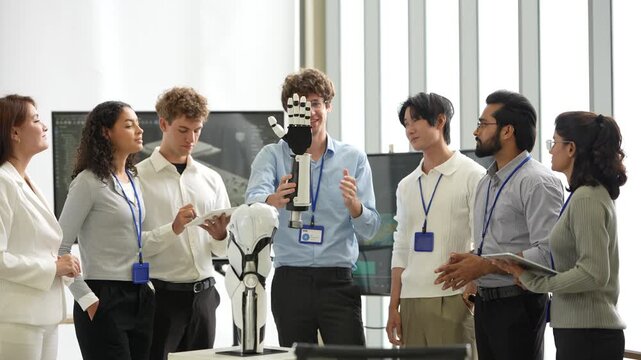 Engineer in blue shirt holds up a bionic robotic hand while colleagues with lanyards and tablets ask questions around a humanoid robot, as the next step is planned, a physical AI concept