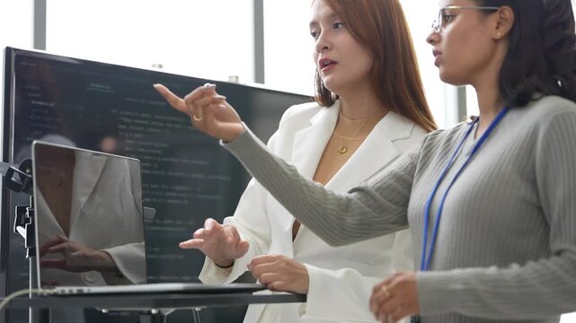 Woman in white blazer explains an idea at a laptop stand while an Indian female colleague with glasses listens and gestures, during an iterative design check, an embodied AI research scene