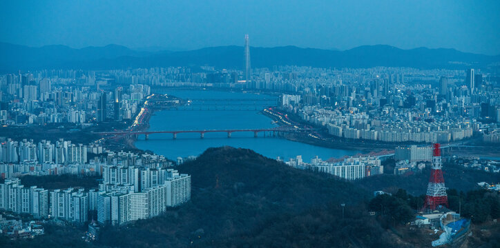 Aerial view of the Han River flowing through the dense urban skyline at dusk, featuring high-rise apartment complexes, bridges, and the distant Lotte World Tower in Seoul, South Korea.
