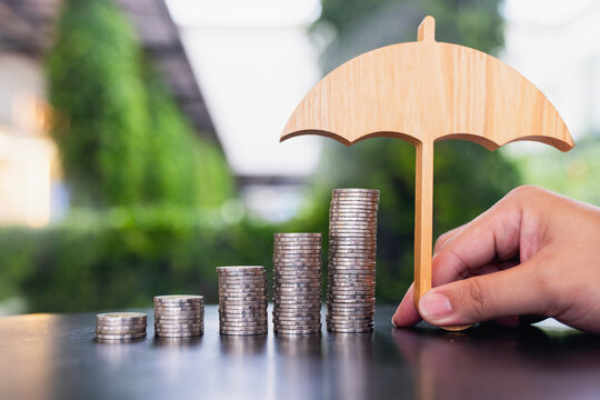 Financial insurance and savings protection concept, hand holding wooden umbrella over ascending stacks of coins on table, symbolizing wealth safeguard, capital investment, and deposit care.