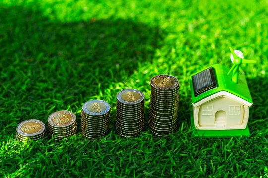 Green energy home and eco housing investment concept, miniature house with solar panel and wind turbine beside rising coin stacks on lawn, symbolizing renewable savings and sustainable value.