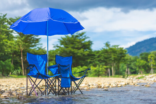 Outdoor camping and nature retreat concept, two blue folding chairs with beach umbrella set up in shallow river stream, representing summer vacation, relaxation, and wilderness travel.