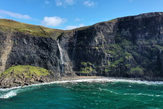 Aerial view of Neist Point waterfall cascading down rugged dark cliffs into the turquoise sea with green grassy slopes in Isle of Skye, Scotland, United Kingdom.