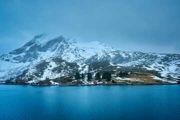 Majestic winter landscape of Lofoten Islands in Norway. Dramatic snow covered mountain towering over a beautiful deep blue sea fjord with a traditional red wooden cabin on the coast. Scenic Arctic nat © bank215