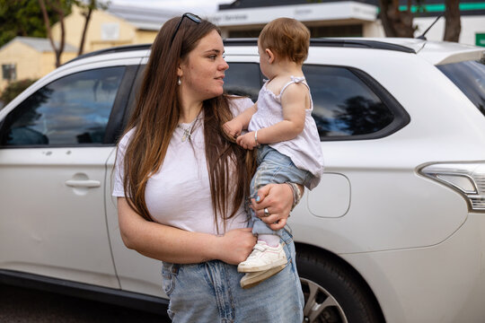 Mother carrying toddler beside family car in suburban neighbourhood