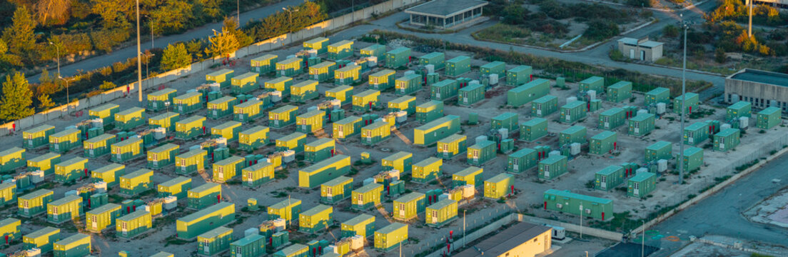 Aerial view of modular battery storage containers and electrical equipment at a modern energy facility, illuminated by warm evening sunlight, Italy.