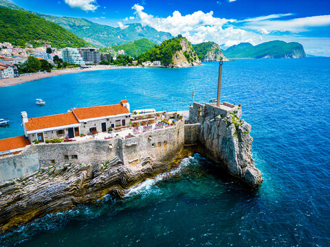 Aerial view of Kastio Castle on a rocky islet with stone walls, orange-tiled roofs, and a tall monument in Petrovac, Budva Municipality, Montenegro.