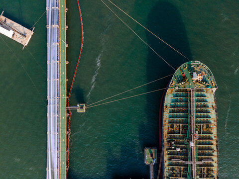Aerial view of a large oil tanker docked at a long industrial pier in the harbor, with calm dark water and mooring lines visible in Cilacap Regency, Central Java, Indonesia.