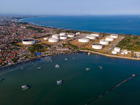 Aerial view of an oil storage terminal with large white cylindrical tanks situated along the coast near a fishing harbor with small boats in Cilacap Regency, Central Java, Indonesia.