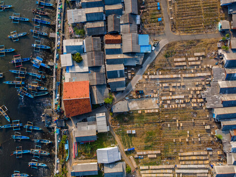 Aerial view of fishing boats docked along a pier next to a coastal village with houses and drying racks in Cilacap Regency, Central Java, Indonesia.
