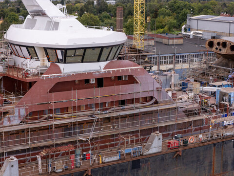 Aerial view of a ship under construction in a shipyard, featuring a white bridge structure, metal hull, scaffolding, and a yellow crane in the background in Gdansk, Poland.