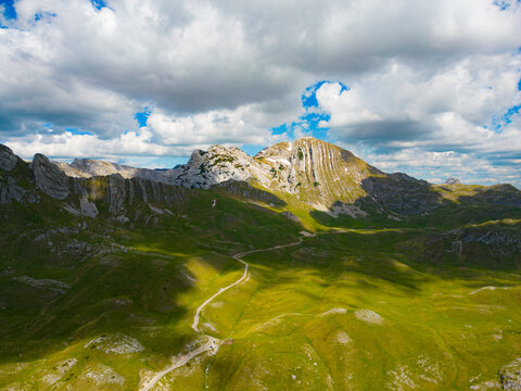 Aerial view of Bobotov Kuk featuring rugged mountain peaks, lush green slopes, and a winding trail under a cloudy blue sky in Zabljak Municipality, Montenegro.