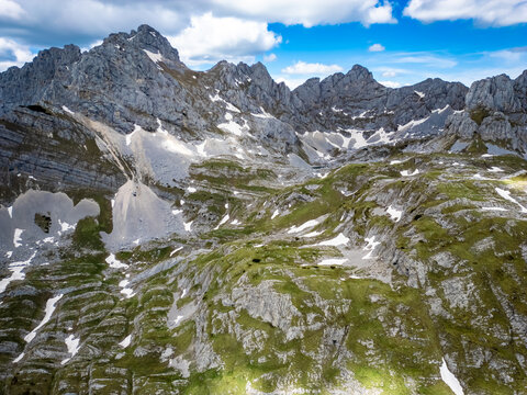 Aerial view of Bobotov Kuk in the Durmitor mountains, featuring rugged rocky peaks, patches of snow, and green slopes under a bright blue sky in Zabljak Municipality, Montenegro.