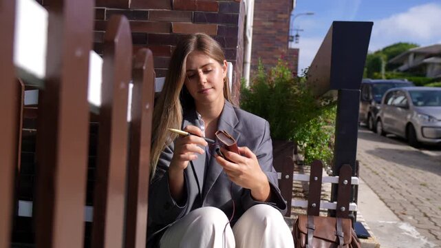 Beautiful young caucasian woman sitting on the front steps of a house on a sunny day, thoughtfully writing her ideas and plans in a personal diary while enjoying the quiet neighborhood street