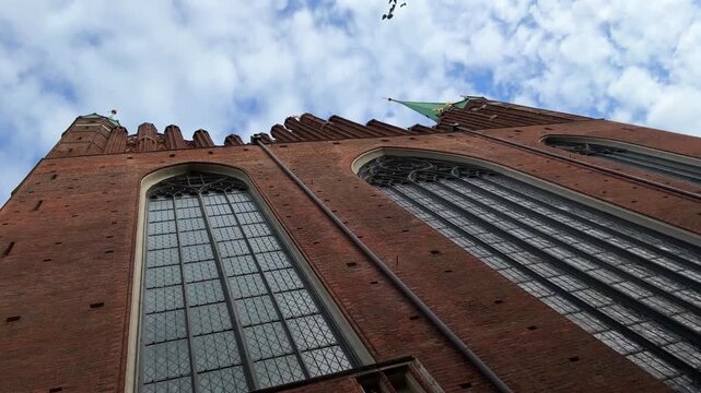 A cinematic horizontal commercial footage looking up and panning along an ancient medieval cathedral wall, showcasing architectural texture, historic masonry work, and green spires against a blue clou