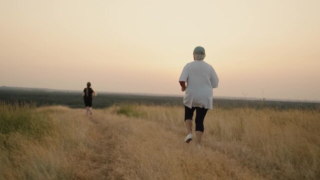 Two women running together along path in tall dry grass during golden sunset.