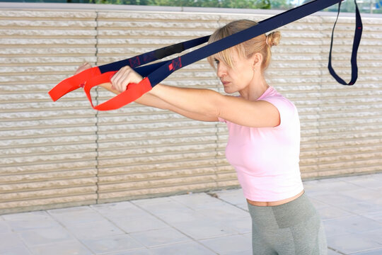Woman performing resistance band chest press outdoors against textured wall in daylight, showing focused posture, toned upper body, balanced stance, and active fitness effort