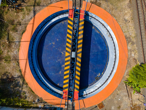 Aerial view of a railway turntable with a blue circular base, an orange outer ring, and a yellow and black striped bridge in Cilacap, Central Java, Indonesia.