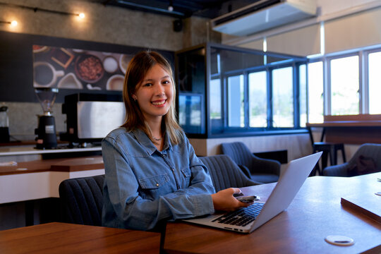 Portrait of smiling young Asian female entrepreneur working on laptop and holding smartphone at cafe table. Gen Z small business owner in modern coffee shop.
