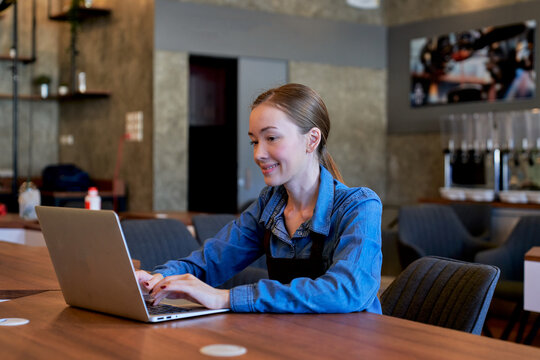 Smiling young female cafe owner typing on laptop at wooden table. Caucasian barista or small business manager checking online orders in modern coffee shop.