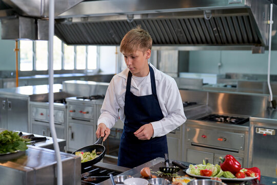 Focused young Caucasian male chef cooking vegetables in frying pan inside commercial restaurant kitchen. Culinary student or apprentice preparing gourmet dish.