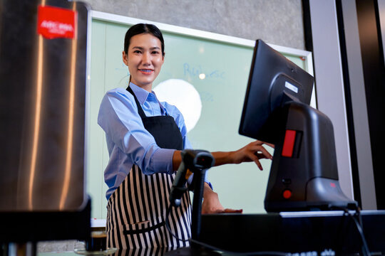 Portrait of smiling young Asian female cashier operating POS cash register terminal in retail store. Friendly small business owner or barista at checkout counter.