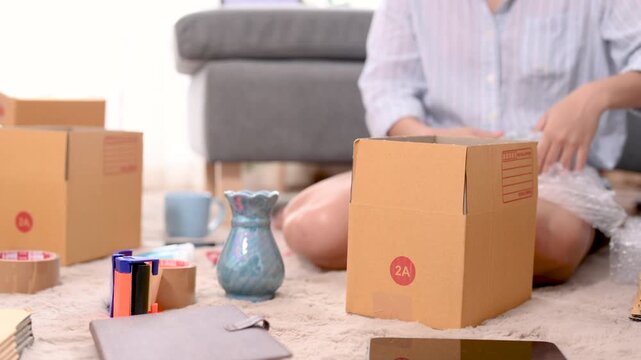 Cardboard boxes of different sizes form a parcel stack inside a home packing area. The cardboard boxes include open and closed brown cartons on the carpet beside a sofa and packing supplies.