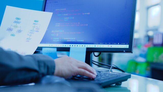 Programmer typing on computer keyboard near monitor displaying script code holding flowchart document diagram paper at workplace desk inside corporate office room.