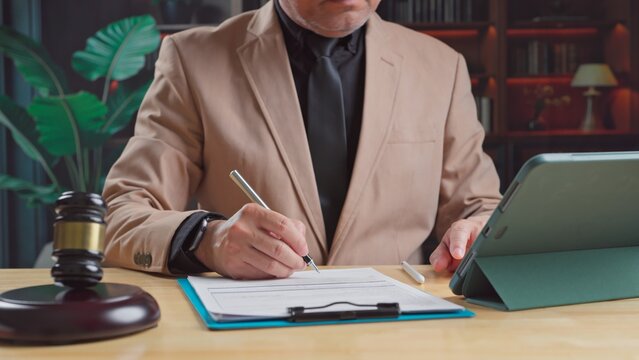 Mature male lawyer sitting at office desk signing legal contract document beside wooden judge gavel digital tablet computer during court case preparation.