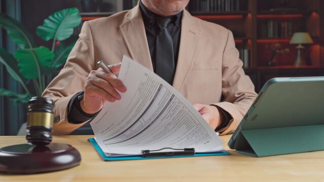 Mature lawyer in formal suit reviewing legal contract papers near wooden gavel digital tablet inside courtroom office workspace.