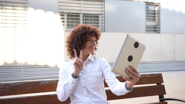 A young woman is sitting on a bench in an outdoor park, using her tablet while enjoying the sun